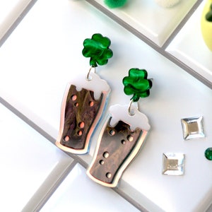 May include: A pair of dangle earrings featuring a green shamrock charm and a brown and white glass beer mug charm.