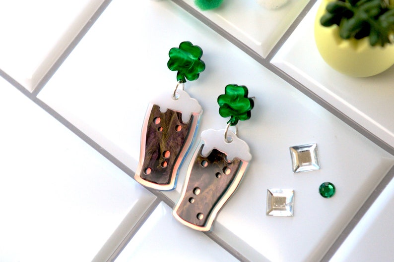 May include: A pair of dangle earrings featuring a green shamrock charm and a brown and white glass beer mug charm.