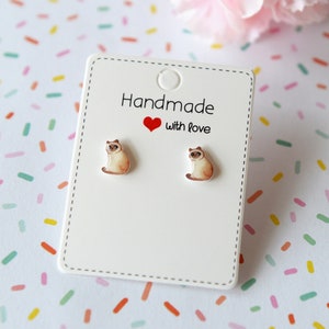 May include: A pair of silver stud earrings featuring a white cat design. The earrings are displayed on a white card with the text "Handmade with love".