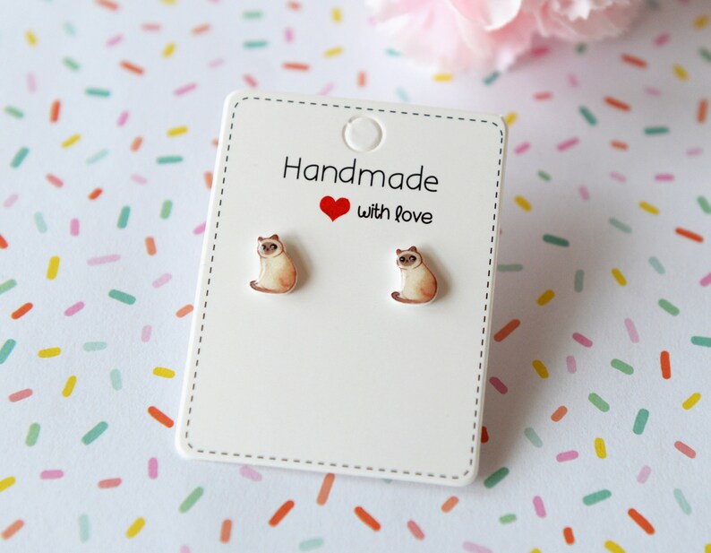 May include: A pair of silver stud earrings featuring a white cat design. The earrings are displayed on a white card with the text "Handmade with love".