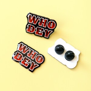 May include: Two black and red enamel pins with the text "WHO DEY" in a striped pattern. The pins are on a yellow background. One pin is shown with the back side, which is white with two black pin backs.