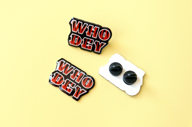 May include: Two black and red enamel pins with the text "WHO DEY" in a striped pattern. The pins are on a yellow background. One pin is shown with the back side, which is white with two black pin backs.