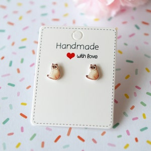 May include: A pair of white card earrings with a small, brown and white cat illustration on each earring. The earrings are on a white card with the text "Handmade with love" in red.