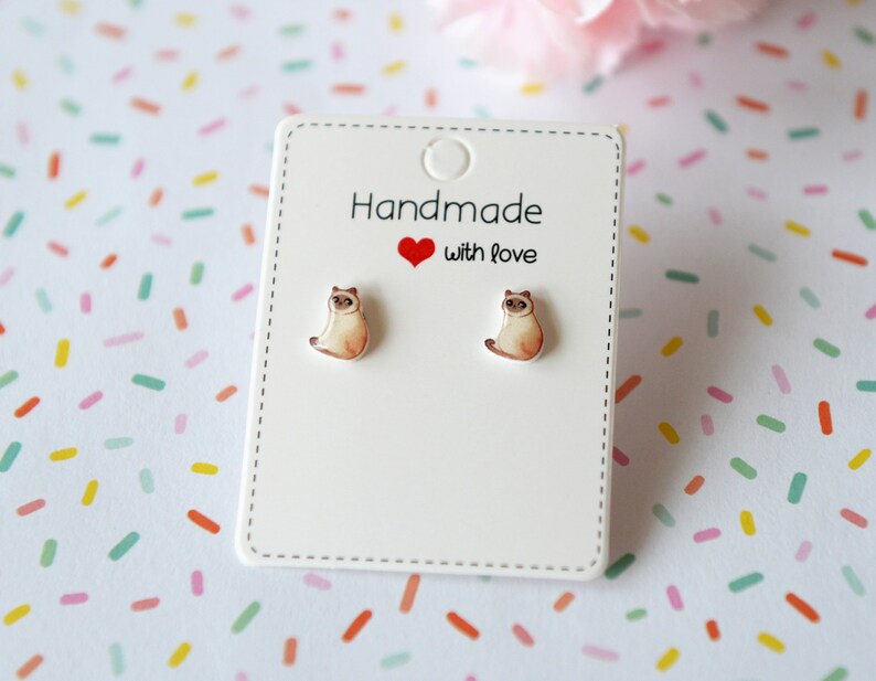 May include: A pair of white card earrings with a small, brown and white cat illustration on each earring. The earrings are on a white card with the text "Handmade with love" in red.