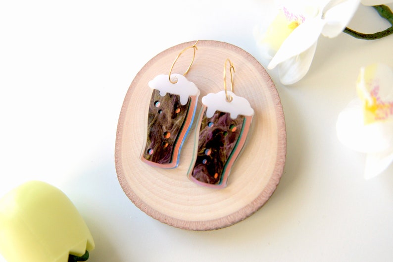 May include: A pair of gold hoop earrings with a colorful, abstract design of a pint glass of beer. The glass is filled with a dark brown liquid and has a white foam head.