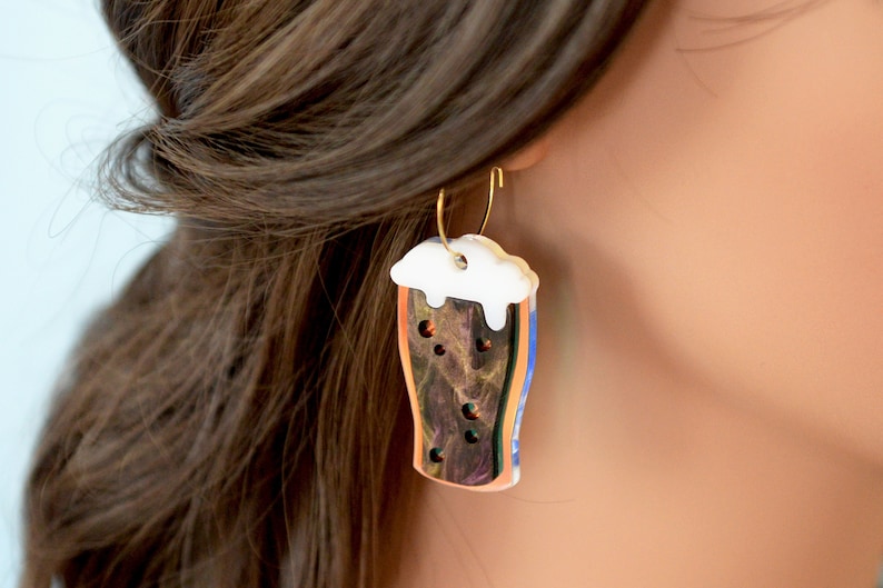 May include: A pair of gold hoop earrings with a glass of beer charm. The charm is made of polymer clay and features a brown beer with a white head. The beer is decorated with a marbled pattern.