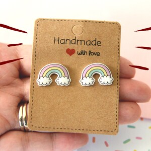K&ouml;nnte beinhalten: Ohrstecker mit Regenbogen-Motiv auf einer braunen Karte. Die Ohrringe zeigen einen bunten Regenbogen mit pastellgelben, blauen und rosa B&ouml;gen, wei&szlig;en Wolken und einer schwarzen Umrandung. Auf der Karte steht "Handmade with love".
