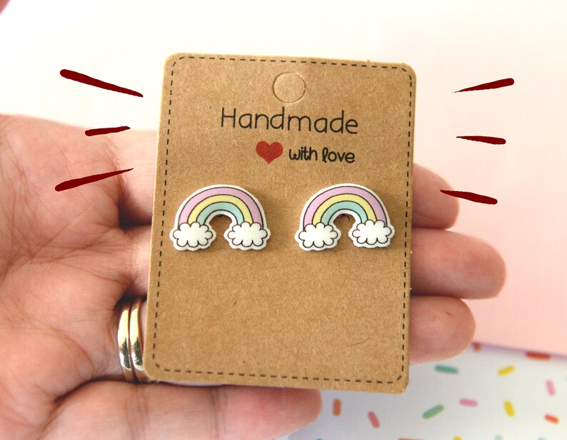 K&ouml;nnte beinhalten: Ohrstecker mit Regenbogen-Motiv auf einer braunen Karte. Die Ohrringe zeigen einen bunten Regenbogen mit pastellgelben, blauen und rosa B&ouml;gen, wei&szlig;en Wolken und einer schwarzen Umrandung. Auf der Karte steht "Handmade with love".