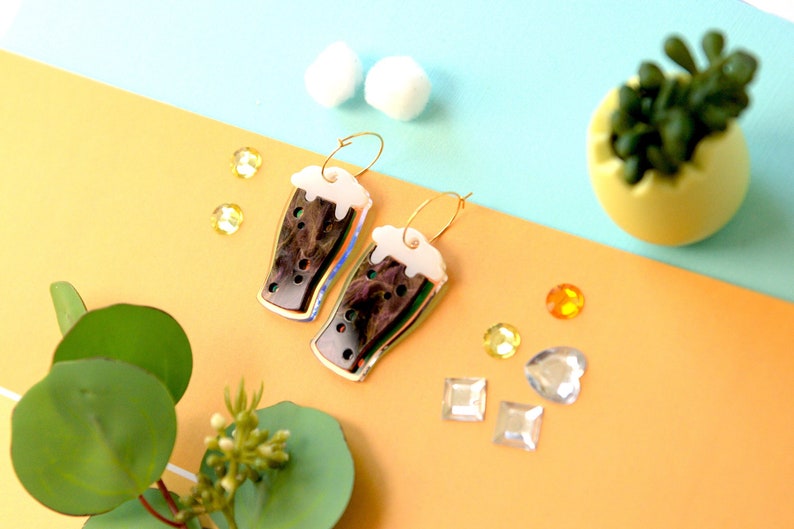 May include: Gold hoop earrings with a dark brown and iridescent glass charm shaped like a pint of beer with a white foam top.