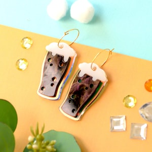 May include: A pair of gold hoop earrings with a black and white beer glass charm. The glass has a white foam top and a black liquid inside. The glass is iridescent and has a gold outline.