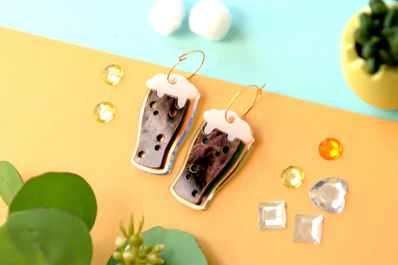 May include: A pair of gold hoop earrings with a black and white beer glass charm. The glass has a white foam top and a black liquid inside. The glass is iridescent and has a gold outline.
