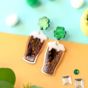 May include: A pair of dangle earrings featuring a green shamrock charm and a glass of dark beer with white foam. The earrings are made of acrylic and have a copper-colored finish.