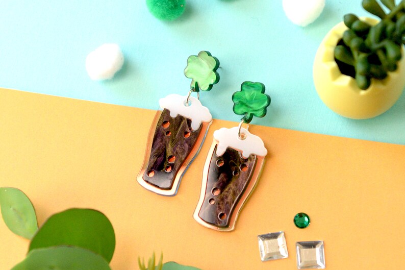 May include: A pair of dangle earrings featuring a green shamrock charm and a glass of dark beer with white foam. The earrings are made of acrylic and have a copper-colored finish.