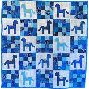 May include: A blue and white patchwork quilt featuring a repeating pattern of stylized horses. The quilt is composed of squares in various shades of blue, including solid, patterned, and dotted fabrics. Each horse has a tan saddle.