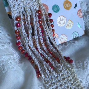 Puede incluir: Un brazalete blanco tejido a crochet con flecos de hilo blanco y cuentas rojas.