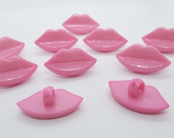 Pode incluir: Uma coleção de dez botões cor-de-rosa em forma de lábios. Os botões são brilhantes e têm um design tridimensional. A parte de trás dos botões tem uma argola para costurar. Os botões são de cor rosa claro.