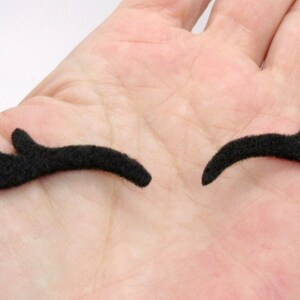 May include: Two black felt eyelashes, each with three points, are shown on a hand. The eyelashes are curved and are meant to be used as a costume accessory.
