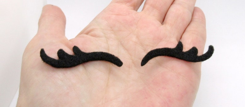 May include: Two black felt eyelashes, each with three points, are shown on a hand. The eyelashes are curved and are meant to be used as a costume accessory.