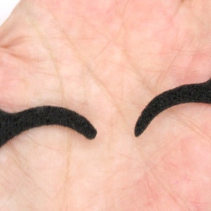 May include: Two black felt eyelashes, one slightly curved and the other with a more dramatic curve, are shown on a hand.