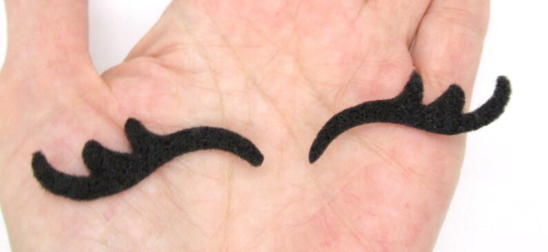 May include: Two black felt eyelashes, one slightly curved and the other with a more dramatic curve, are shown on a hand.