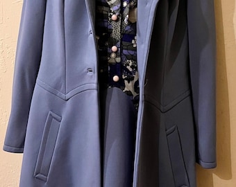 May include: A periwinkle blue coat with wide lapels and two front pockets, worn over a dark blue and white floral patterned blouse featuring a pussy-bow neckline and light pink buttons down the center front, displayed on a mannequin.