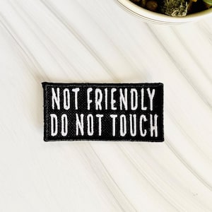 May include: Black iron-on patch with white text that reads "Not Friendly Do Not Touch".