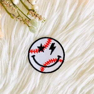 May include: A white baseball with red stitching and black lightning bolt eyes, forming a smiley face. The patch is on a white fluffy surface.
