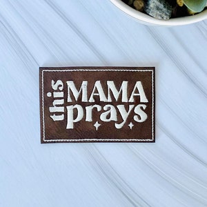 May include: A brown leather patch with white embroidery that reads "this mama prays".