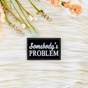 May include: A black patch with white text that reads "Somebody's PROBLEM" on a white fluffy background with a sprig of pink flowers.