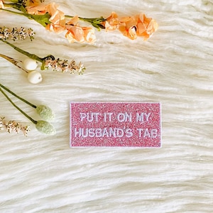 May include: A pink glitter patch with the text "PUT IT ON MY HUSBAND'S TAB" in white lettering. The patch is on a white fuzzy surface with a bouquet of peach-colored flowers.