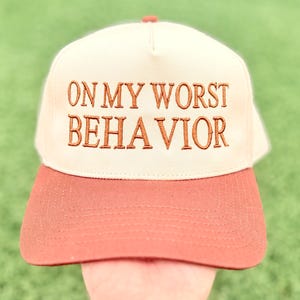 May include: A cream-colored baseball cap with a structured front panel and a rust-orange brim. The front of the cap features the embroidered text ON MY WORST BEHAVIOR in a dark brown, serif font, centered across the crown of the hat.