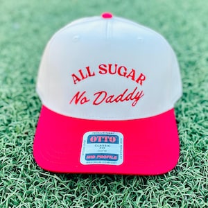 May include: A white and hot pink baseball cap with the text "ALL SUGAR No Daddy" embroidered in red. The cap has a classic fit and a mid-profile design. The cap is sitting on a green surface.