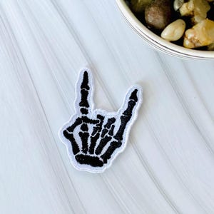 Peut inclure: Écusson brodé noir et blanc en forme de main de squelette faisant le signe des cornes. Le patch a une bordure blanche et est posé sur un fond blanc texturé. Un petit pot de plantes est visible en haut à droite.