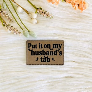 May include: A rectangular, tan patch with the phrase "Put it on my husband's tab" in black lettering. The patch is decorated with two small star symbols. The patch is on a white, fluffy surface, with peach-colored flowers in the background.
