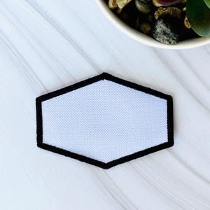 May include: A hexagonal patch featuring a solid white center with a textured fabric weave, framed by a thick, solid black embroidered border. The patch is positioned centrally, showcasing a clean, geometric shape with sharp, defined edges.
