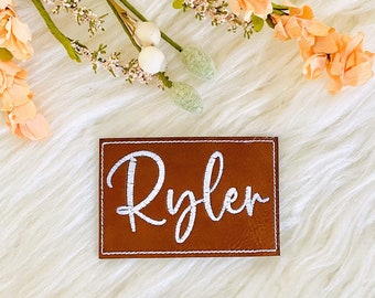 May include: A rectangular brown leather patch featuring the name Ryler embroidered in white script lettering. The patch has a white stitched border around the perimeter, providing a clean and defined edge to the brown material.