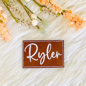 May include: A brown rectangular patch with the name "Ryler" embroidered in white script. The patch has a white border and is set against a white, fluffy background. Peach-coloured flowers and greenery are in the background.