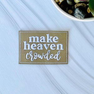 May include: A brown leather patch with white embroidery that reads "make heaven crowded".