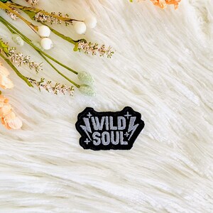 May include: A black embroidered patch with white stitching that reads "WILD SOUL" with lightning bolts on either side. The patch is on a white fluffy surface.