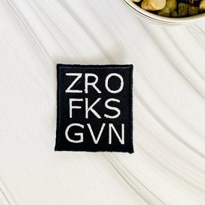 May include: A black square patch with white embroidered text reading "ZRO FKS GVN." The patch is centered on a white and gray surface, with a small potted plant in the background.
