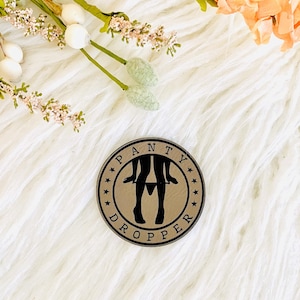 May include: A gold-colored round pin with a black silhouette of a person with their pants falling down. The pin says "Panty Dropper" around the edge.