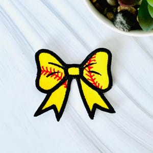May include: A yellow softball bow with black trim and red stitching details. The bow is a decorative accessory, likely for hair or clothing, and is set against a white background with a small plant in the background.