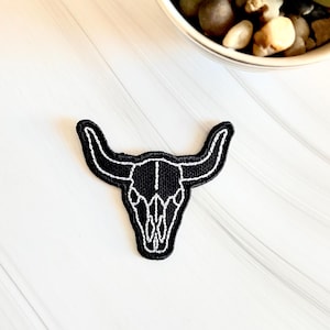 May include: A black embroidered patch featuring a white outline of a bull skull. The patch has a black background and a white stitched border. The design is a western-themed accessory.