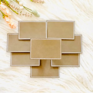 May include: A collection of rectangular, tan-colored name tags with silver trim, arranged in a starburst pattern. The tags are on a fluffy, white surface, with a floral arrangement in the upper left corner.