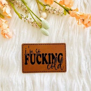 May include: A brown leather patch with the text "I'm so FUCKING cold" in black lettering.