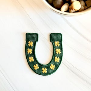 May include: A green embroidered horseshoe patch with gold shamrocks. The patch is in the shape of a horseshoe and features a row of gold shamrocks along the inside edge. The patch is likely for St. Patrick's Day or general good luck.