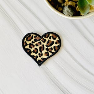 May include: Embroidered heart-shaped patch with a leopard print design. The patch features a black border and a tan and brown animal print pattern. The patch is likely for clothing or accessories.