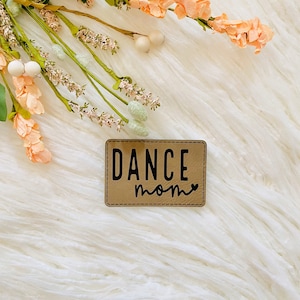 May include: A brown leather patch with the text "DANCE MOM" in black lettering and a small heart symbol.