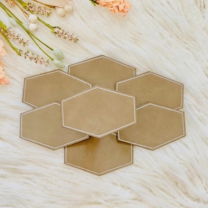 May include: Set of six hexagonal coasters in a light brown color with white stitching around the edges. The coasters are arranged in a honeycomb pattern.