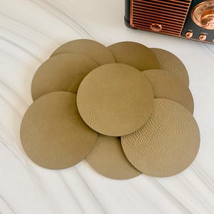 May include: A collection of circular, tan-colored coasters with a textured surface. The coasters are arranged in an overlapping stack. A vintage-style radio with a dark gray casing and copper-colored speaker grille is positioned above the coasters.